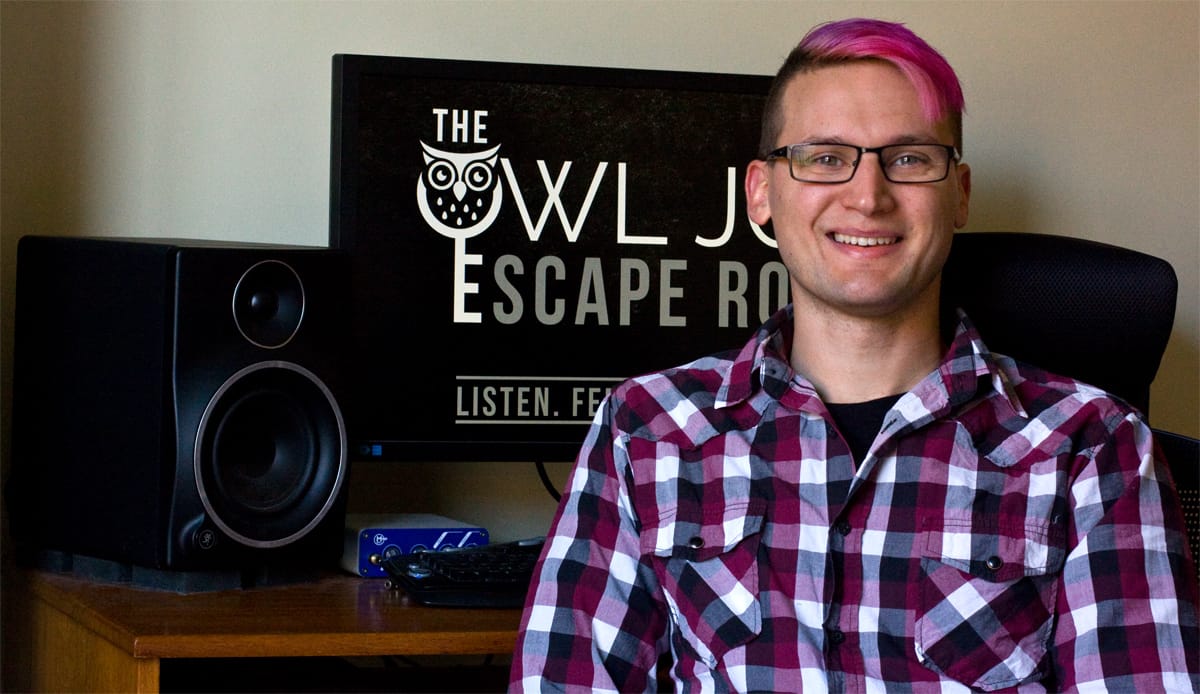 A man with pink hair and a plaid shirt sits in front of his work station with The Owl Job on the screen.