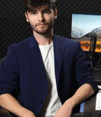 A man with a shaggy beard and hair in a blazer sits at his workspace.