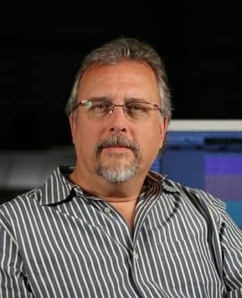 A man with gray hair and a beard sits at his workstation.