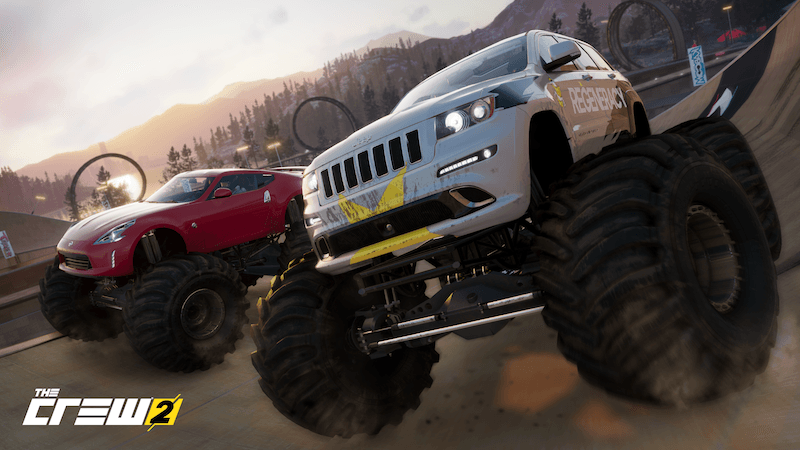 Two monster trucks race on a highway along a mountainous shore.