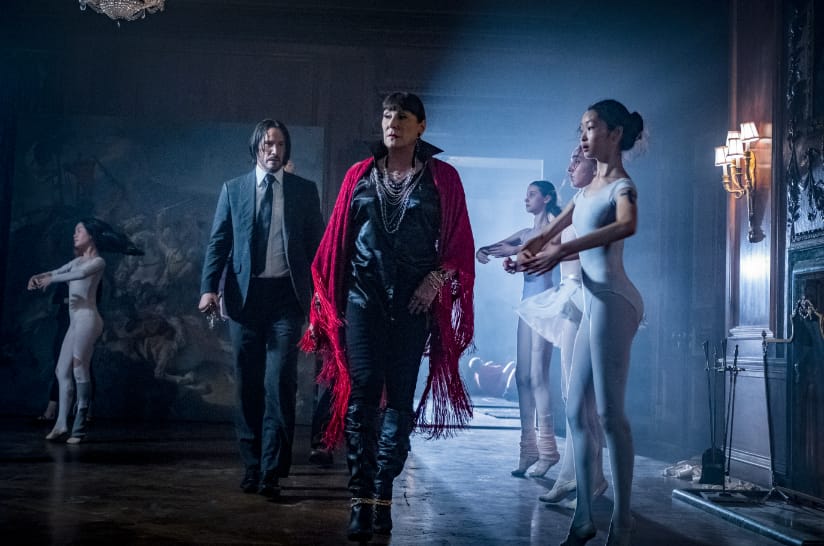 John Wick walks behind a women in black with a red shawl in front of ballet dancers