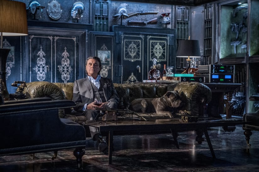 An older man (Ian McShane) sits with a large brown dog in an dark, exquisitely designed room