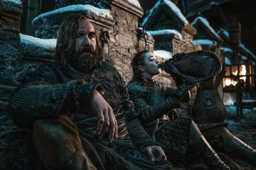 The Hound and Arya lean back against a wall at night and drink.