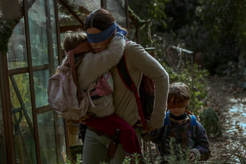 A woman carries her children through a garden as they wear blindfolds.