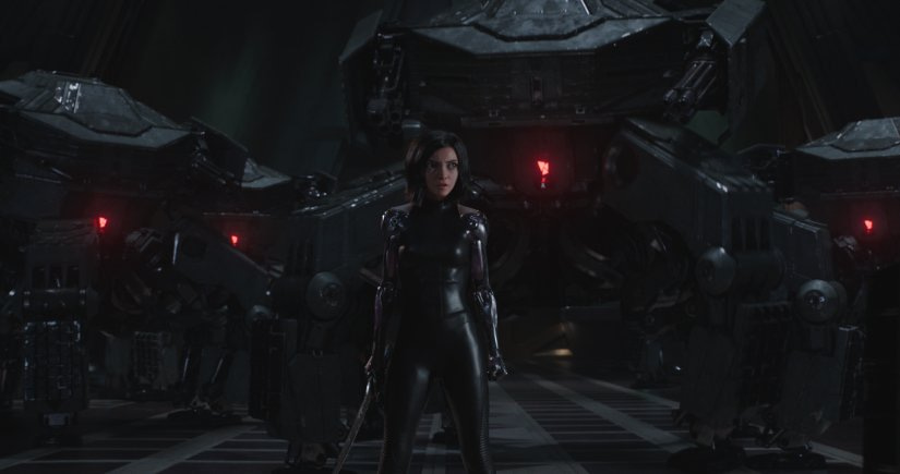 Alita, in all black, stands in front of massive dark machinery.