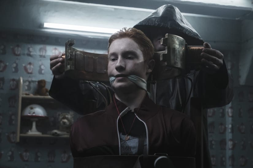 A red-headed teenager is gagged and placed in a device by a hooded man.