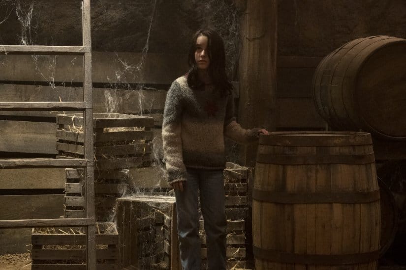 A girl stands in the cob-webbed cellar next to a barrel.
