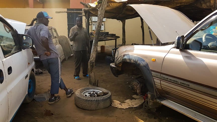 Two mechanics talk in their garage next to three cars in need of repair.