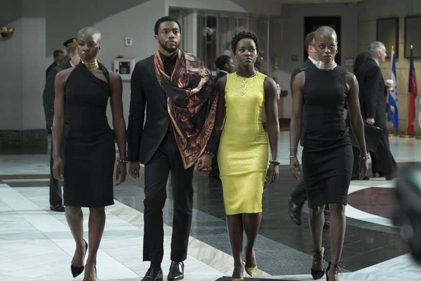 Black Panther wears a black suit and scarf and three Dora Milaje walk in sleek dresses