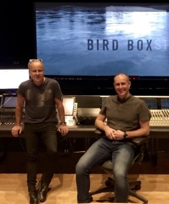 Two men smile as they stand and sit near their soundboard.