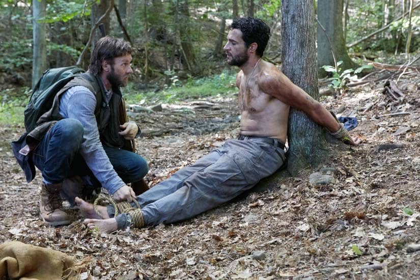 A man kneels, holding the fastened feet of another man whose hands are tied behind his back to a tree
