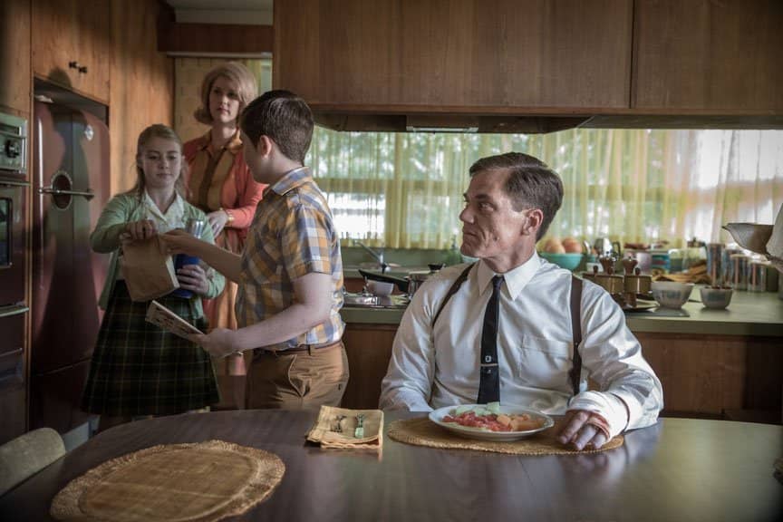 A stern man sits at a table and looks back at his family in the kitchen.